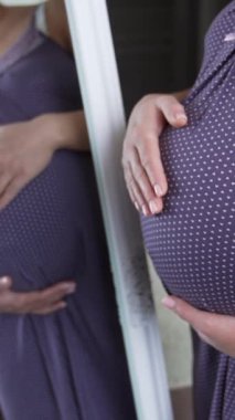 Beyaz bir kadının karnını aynanın önünde okşamasını beklemek. Hamile kadın doğum için hazırlanıyor. Müstakbel anne rahat yatak odasında pijamayla bekliyor. Dikey video