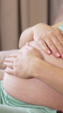 Müstakbel annesinin koltukta dinlenirken koca karnını okşaması. Bir kadın koca karnında çocuğunu okşuyor. Arka planda iki küçük bebek çorabı var. Dikey video