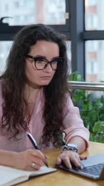 İş kadını masada dizüstü bilgisayarla çalışıyor. Bilgisayarda çalışıp notlar alıyorum. Kadın işine odaklanmış. Dikey video