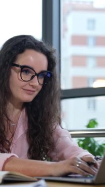 Bilgisayarda çalışan ve gülümseyen güzel bir kadın. Keyfi yerinde bir kadın işçi ofiste çalışır. Geniş pencereli büyük ofisteki işyerinde. Dikey video