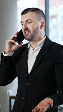 İş adamı cep telefonuyla güzel bir konuşma yapıyor. Kendine güvenen adamın telefonda iş konuşması var. Arka planda büyük pencereler var. Dikey video