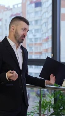 Takım elbiseli bir iş adamı konferans masasında oturan iş arkadaşlarıyla konuşuyor. Erkek lider elinde dosyayla iş arkadaşlarına bir dosya gösteriyor. Dikey video