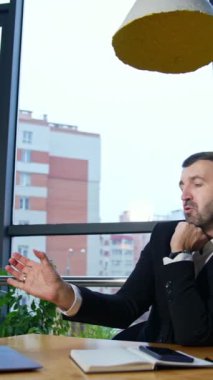 Bilgisayarda iş görüşmesi. Orta yaşlı girişimci büyük panoramik pencerelerin önündeki masada oturur. Arka planda şehir manzarası. Dikey video