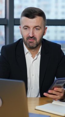 Ceketli ve takım elbiseli bir adam laptopta biriyle konuşuyor ve elinde akıllı telefon tutuyor. Ofisteki ortaklara video çağrısı. Görüşmeler dizüstü bilgisayarla yapılıyor. Dikey video