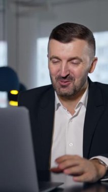Bilgisayarla iş görüşmesi. İş adamı dizüstü bilgisayarın önünde konuşurken konuşuyor ve gülümsüyor. Arka planda bulanık bir ofis. Dikey video