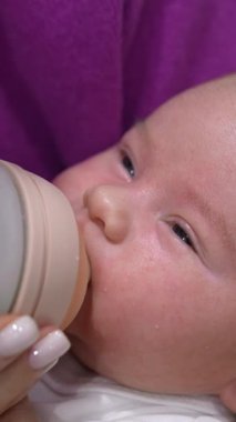 Yeni doğmuş bebek, annesinin elinde duran bir şişeden beslendi. Bebek çocuk beslenirken gözlerini ışığa doğru çeviriyor. Kapatın. Dikey video