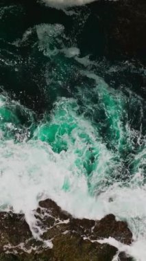 Aquamarine suyu okyanustan süzülen engebeli kayaları eziyor. Yosun kümesi kayanın etrafında toplanıyor. Üst Manzara. Dikey video