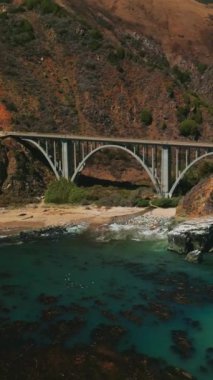 California dağlarındaki kemerli köprü. Arabalar hareket halinde. Güneşli bir günde Pasifik Okyanusu kıyısından insansız hava aracı görüntüleri yükseliyor. Dikey video