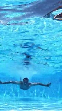 Yüzen mutlu bir adamın sualtı görüntüsü. Adam havuzda suyun altında yüzüyor ve ellerini kameraya doğru gösteriyor. Dikey video