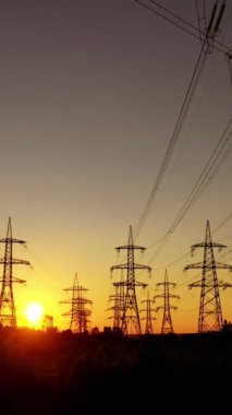 Turuncu gün batımına karşı elektrik kuleleri. Akşamları yüksek voltajlı elektrik hatları. Elektrik direğinin siluetleri. Dikey video