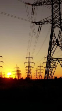 Gün batımında kulelerde elektrik iletimi. Yüksek voltajlı elektrik hatları. Turuncu gökyüzü arka planında elektrik direkleri. İHA görüntüsü. Kamera yükseliyor. Dikey video