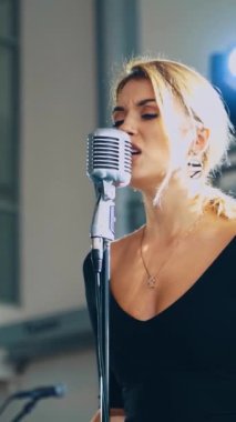 Genç ve güzel bir şarkıcının portresi. Sarışın kadın sahnede duruyor ve mikrofona şarkı söylüyor. Ağır çekim. Dikey video