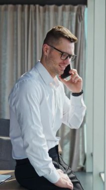 Ofiste resmi kıyafetler ve gözlük takan mutlu adam. İş adamı telefonda konuşuyor, pencereye bakıyor. Dikey video