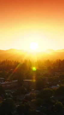 Güneşin batışının ışıl ışıl ışıl ışılında güzel, rahat yeşil bir şehir. Güneş dağların arkasında saklanıyor. Arkaplanda turuncu gökyüzü. Hava görüntüsü. Dikey video