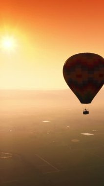 Turuncu gökyüzünde süzülen renkli sıcak hava balonu. Aerostat gün batımında vadinin üzerinde uçuyor. Üst Manzara. Dikey video