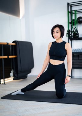 Siyah spor elbiseli bir kadın spor salonunda bacağını yoga minderine uzatıyor..
