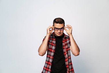 Kırmızı ekose gömlekli ve siyah tişörtlü bir adam gözlüğünü iki eliyle ayarlar, hafifçe aşağı bakar..