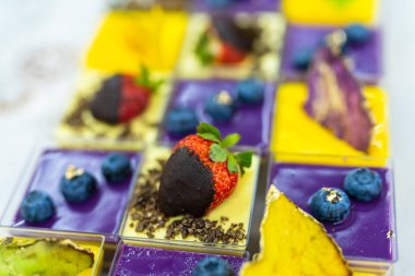 Çilek ve yaban mersini gibi çeşitli meyvelerle kaplı küçük kare tatlıların yanı sıra çikolata ve altın yaprak..