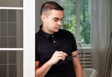 Siyah polo tişörtlü bir adam elinde gözlükleriyle pencerenin yanında duruyor. Gözlüklere bakıyor..