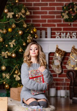 Gri kazaklı bir kadın elinde Noel hediyesiyle şöminenin yanında oturuyor. Solda bir ağaç ve mantoda çoraplar var..