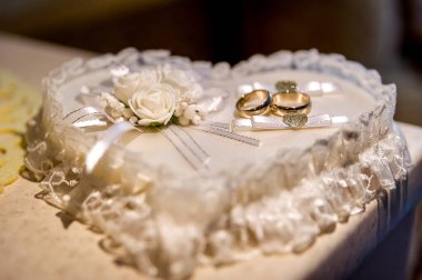 İki altın alyans, beyaz çiçekler ve dantellerle süslenmiş kalp şeklinde bir yastığın üzerinde duruyor..