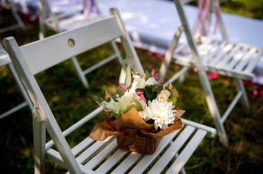 Beyaz katlanır bir sandalye çimenli bir açık alanda, kahverengi kağıda sarılmış bir buket beyaz ve pembe çiçekle koltukta oturur..