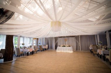 Tavanı ve duvarları beyaz kumaşla kaplayan bir düğün yeri. Masalar ve sandalyeler odanın etrafına kurulmuş, ortada bir baş masa var..