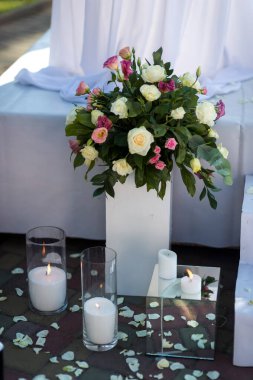 Beyaz ve pembe güller ve diğer çiçekler, yere saçılmış mumlar ve çiçek yapraklarıyla beyaz bir vazoda sergilenir..