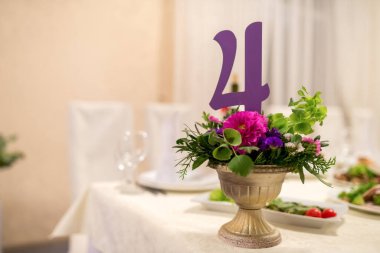 Mor bir 24 numara, çiçekli bir vazonun üzerinde, beyaz bir masa örtüsünün üzerine yerleştirilmiş altın bir vazoda duruyor..