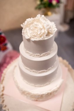 Üzerinde büyük beyaz bir çiçek bulunan üç katlı beyaz bir düğün pastası. Pasta pembe bir masa örtüsü ve altın bir tabakta..