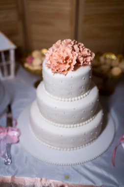 Üzerinde pembe bir çiçek olan üç katlı bir düğün pastası, beyaz krema ve inci aksanıyla süslenmiş, beyaz bir masa örtüsünün üzerinde oturuyor..