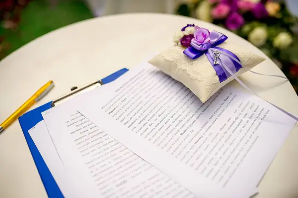 Stock image A white table with wedding ceremony documents and a ring pillow with purple flowers and a ribbon.