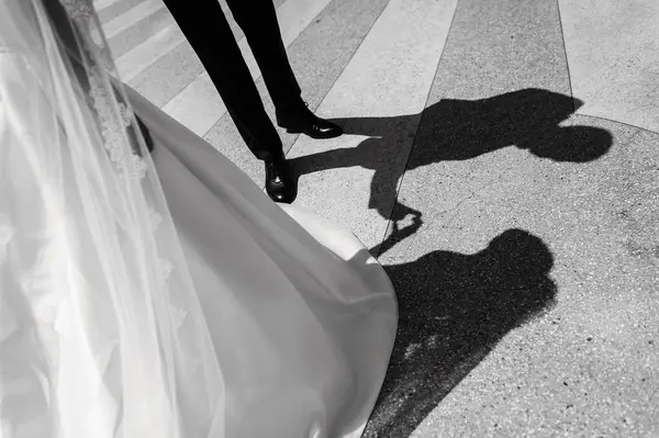 stock image A bride's dress and a groom's legs are visible in the foreground, with their shadows cast on the pavement behind them.