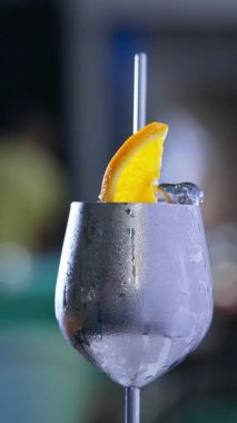 Sisli yüzeyi olan dönen metal şarap kadehleri. Buzlu ve portakal dilimli kokteyl ve içinde metal pipet. Kapatın. Arkaplan bulanık. Dikey video