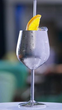 Misted metal şarap kadehi ve dönen masanın üzerinde metal pipet. Üstünde buz küpleri ve portakal dilimleri olan kokteyl. Kapatın. Arkaplan bulanık. Dikey video