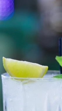 Bardağın üst kısmı hafif içecekle dolu ve iki siyah pipet var. Buz küpleri, limon dilimi ve üstüne nane yaprağı. Kapatın. Arkaplan bulanık. Dikey video