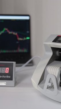 Adam 100 dolarlık banknotları bir para sayma makinesine koyuyor. Amerikan banknotları ekipman olarak sayıldı. Arkaplanı bulanık olan bir dizüstü bilgisayar. Dikey video