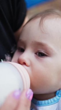 Dışarıda bankta oturan annesinin kucağında tatlı bir bebek. Küçük çocuk şişeyi emziriyor ve ayaklarına dokunuyor. Yavaş yavaş mesafeli yaklaş. Dikey video