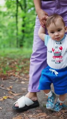 Küçük oğlana yürümeyi öğretiyorum. Anne bebeğini ellerinden tutuyor ve çocuğuna parkta yol boyunca önderlik ediyor. Doğa bulanık zemin. Dikey video