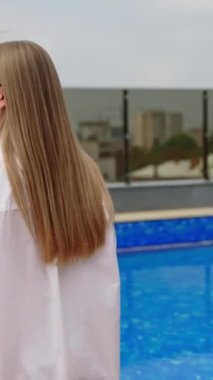 Beyaz mayo ve gömlekli genç sarışın bayan yüzme havuzunun yanında yürüyor. Binanın çatısındaki dinlenme yeri. Şehir manzarası arka planda. Dikey video