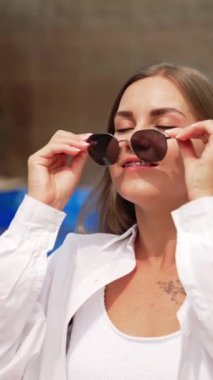 Havuzun yanında oturan beyaz gömlekli mutlu, rahat genç bayan. Kadın güneş gözlüğünü takar ve kameraya gülümser. Dikey video