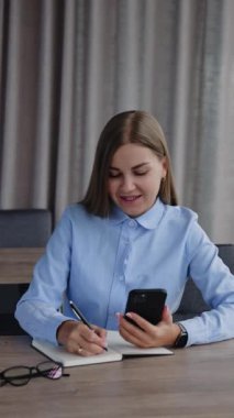 Resmi kıyafetli sarışın kadın ofiste çalışıyor. Uzun saçlı bayan telefonundan kağıt defterine not alıyor. Dikey video