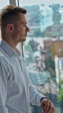 Ofisin penceresinin yanında beyaz gömlekli, kendine güvenen bir iş adamı. Erkek gözlüğü takar ve kameraya gülümser. Dikey video