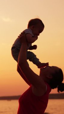 Mutlu anne gün batımında nehrin kenarında bebeğiyle oynuyor. Kadın oğlunu havaya kaldırır ve çocuk yüzüne küçük ayaklar koyar. Dikey video