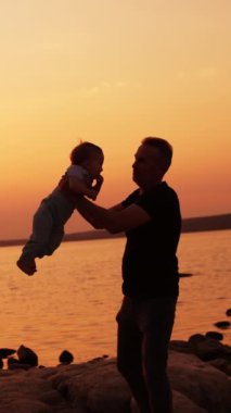 Babası küçük oğluyla nehir kıyısında fırıl fırıl dönüyor. Günbatımında doğada geçirilen aile zamanı. Arkaplanda pembe gökyüzü. Dikey video