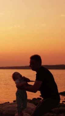 Babam küçük oğlunu nehrin kenarında eğlendiriyordu. Baba sallanıyor, oğlunu savuruyor ve çocuğu mutlu. Günbatımı arka planı. Dikey video