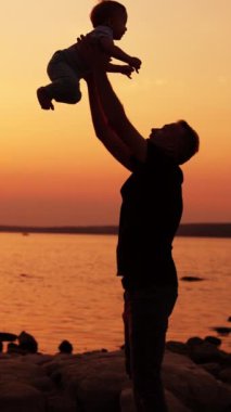 Nehir kıyısında bebeğiyle oynarken dikkatli ol. Sevgi dolu bir babanın oğlunu ve çocuğunu terk etmesi sevinç verici. Arka planda güneşin batışını. Dikey video