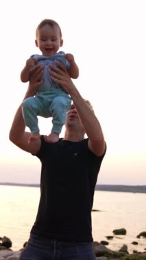 Sevimli mutlu erkek bebek babası tarafından havaya atıldı. Baba ve oğul gün batımında nehir kenarında iyi vakit geçiriyorlar. Dikey video