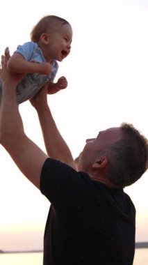 Babacığın güzel çocuğunu havaya fırlatıyor. Yazın nehir kenarında ailecek vakit geçiririz. Arka planda bitki silueti. Dikey video