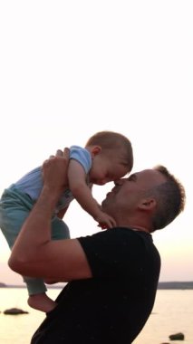 Sevgi dolu bir baba oğluyla konuşuyor ve onu öpüyor. Gülen çocuk babasının göğsüne çıplak ayakla basıyor. Arka planda nehir var. Dikey video